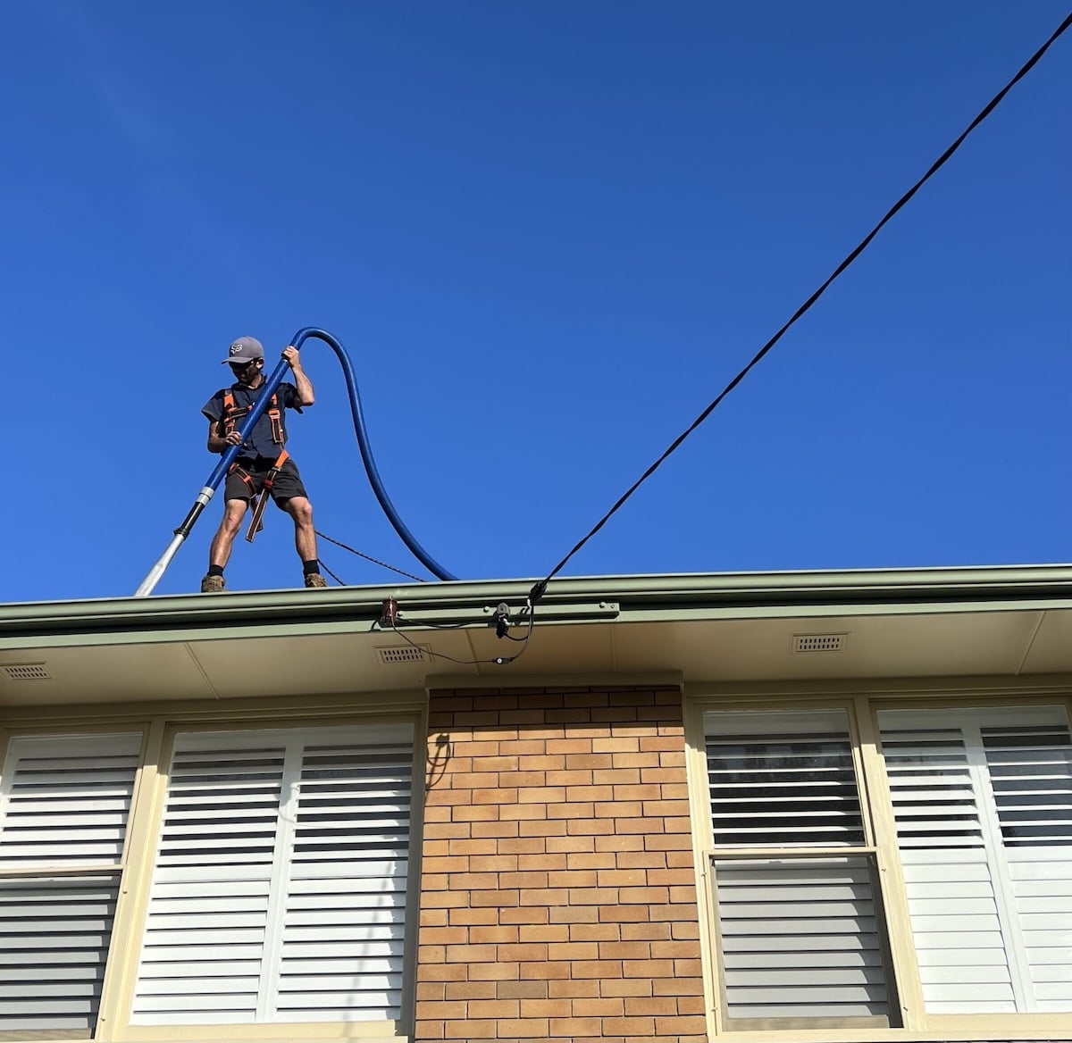 A cropped, square-format shot of a worker in a safety harness standing on the gutter line of a brick house, handling a large blue gutter-cleaning vacuum hose.