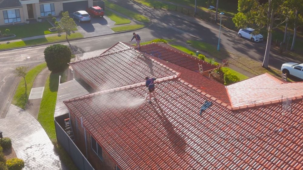 Two workers roof cleaning a large residential house, using pressure washers to spray mist onto the orange terracotta tiled roof.