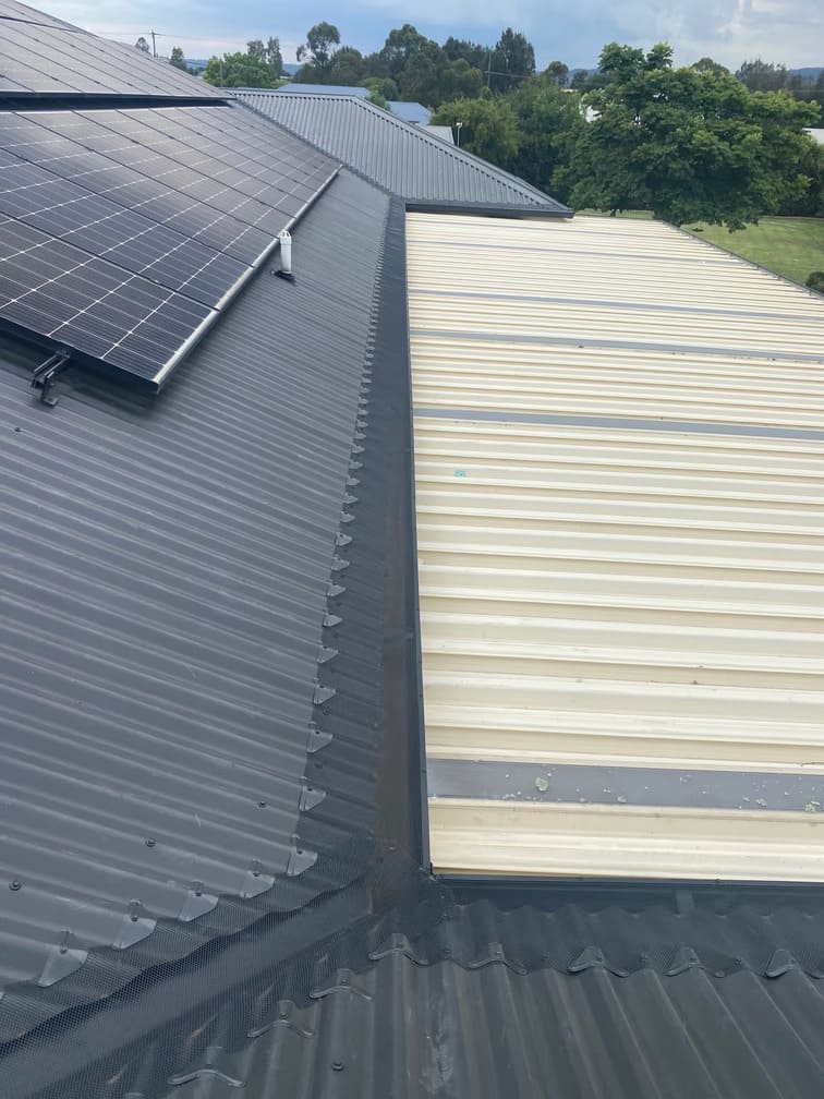 A box gutter valley where a dark grey corrugated metal roof with solar panels meets a cream-coloured patio roof, protected with mesh gutter guards.