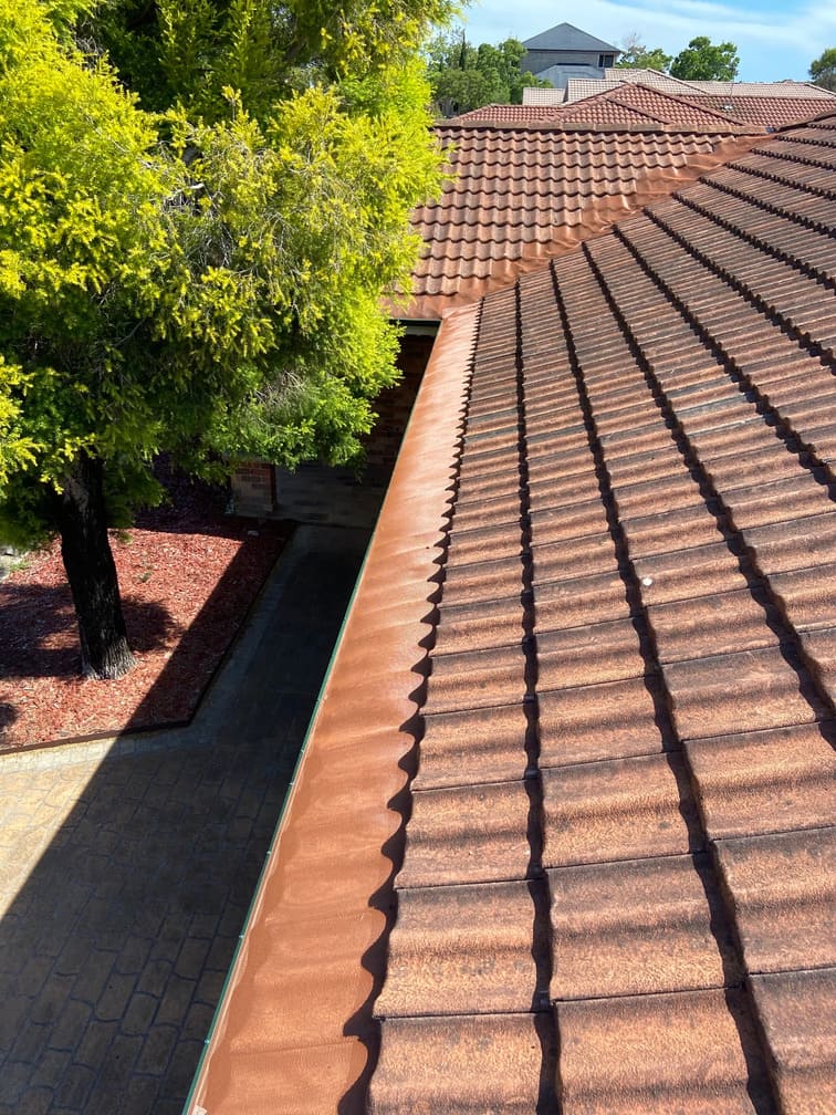 A brown tiled roof, featuring a clean, newly installed matching brown valley gutter guard system, with a green tree and walkway visible below.